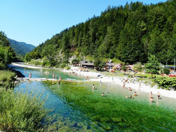 Das Wasser ist klar, blau grün. Menschen gehen über eine Schotterbank in den Fluss. Im Hintergrund Hügel, Wälder und Holzhäuschen.