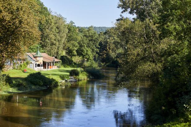 Grün-braunes Wasser, Bäume, eine Liegewiese und im Hintergrund ein Holzgebäude, das einst das Flussbad beherbergte.