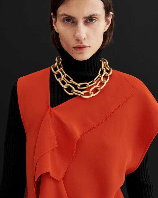Eine Frau mit einem schwarzen Rollkragenpullover, einer orangefarbenen Bluse und einer goldenen Halskette.