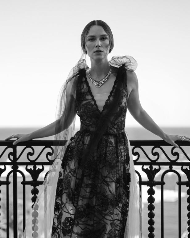 Keira Knightley posiert in einem schwarzen Kleid mit Spitze auf einem Balkon am Meer.