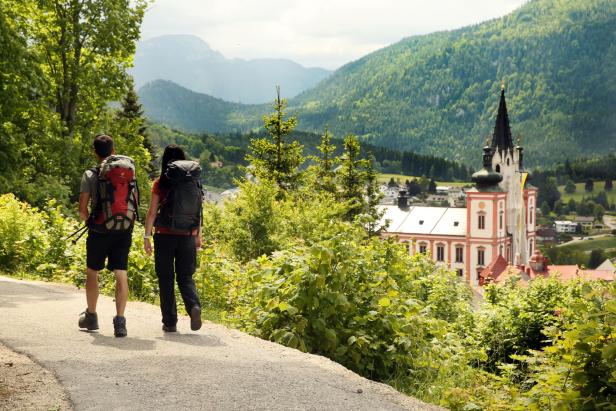 Zwei Wanderer auf einem Weg mit Blick auf eine Kirche und bewaldete Hügel.