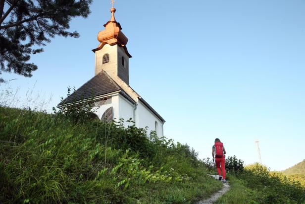 Eine Wanderin geht auf einem Pfad in Richtung einer kleinen Kirche.