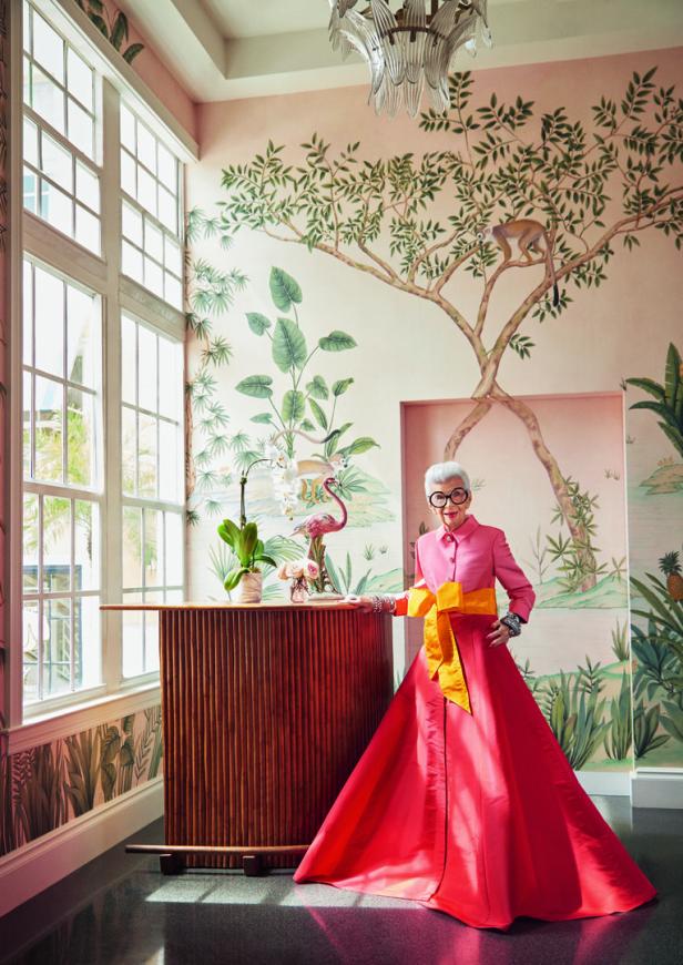 Iris Apfel posiert in einem farbenfrohen Kleid vor einer Wand mit Dschungelmotiven.