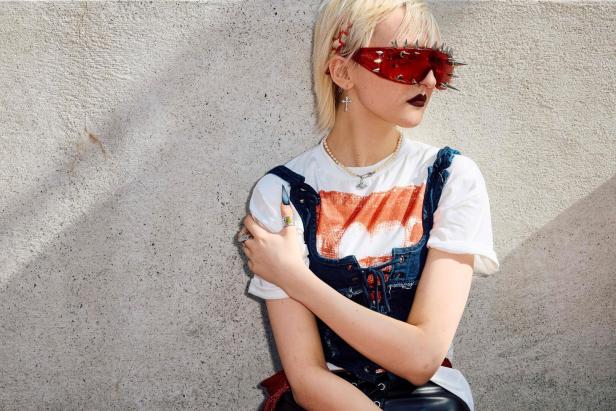 Eine Frau mit blonden Haaren und Nietensonnenbrille lehnt an einer Wand.