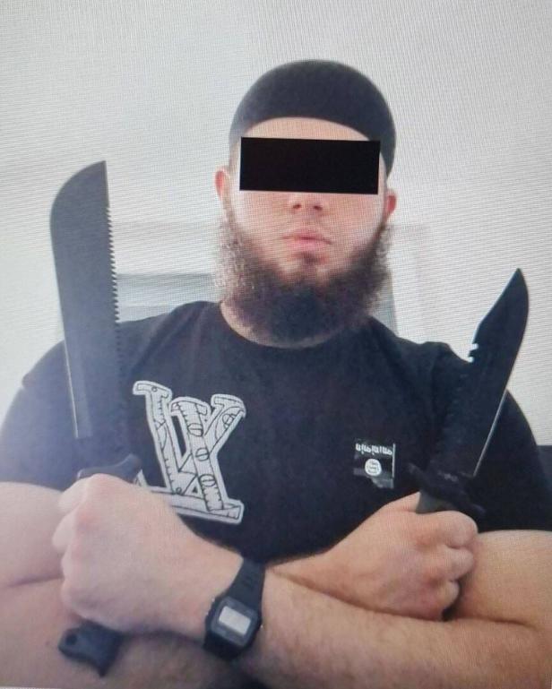 Ein Mann mit Bart und Mütze hält zwei große Messer vor der Brust.