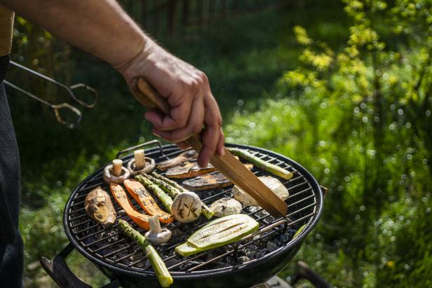 Kleiner Griller, auf dem Rost liegen Zucchini, Champignons, Spargel und Melanzani, ein Mann wendet das Gemüse mit einer Grillzange.
