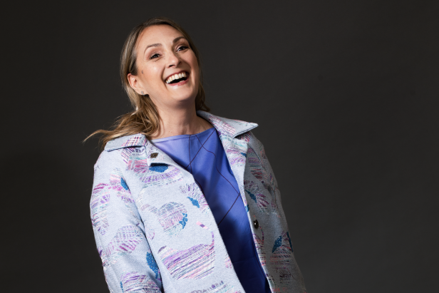 Eine lachende Frau mit blonden Haaren trägt eine blaue Jacke mit Muster.