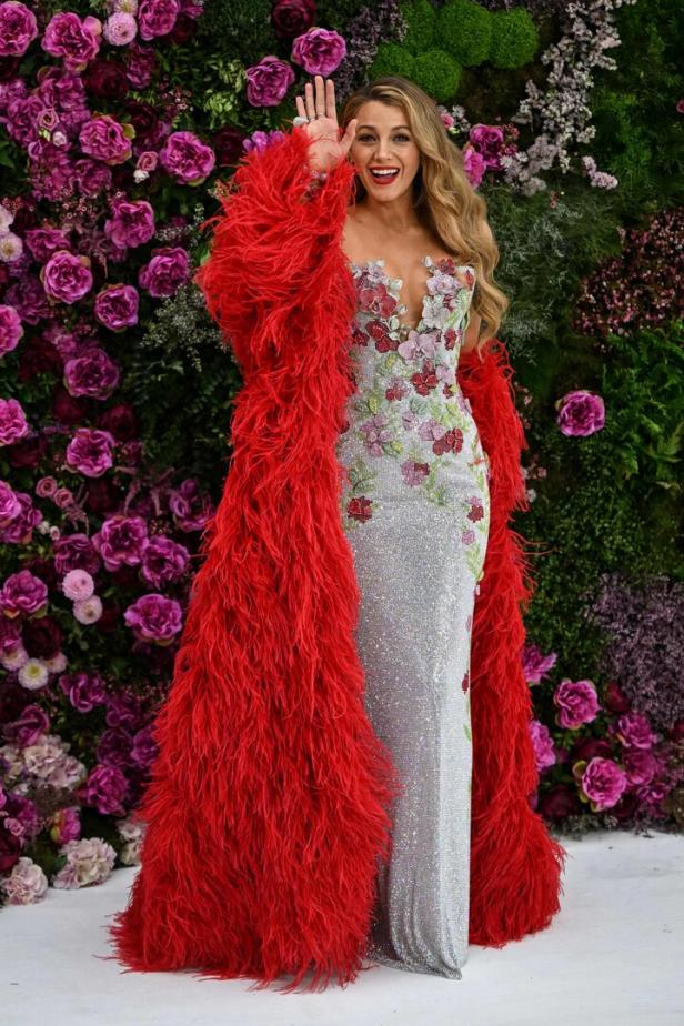 Blake Lively in einem silbernen Kleid mit Blumenapplikationen und einem roten Federboa vor einer Blumenwand.