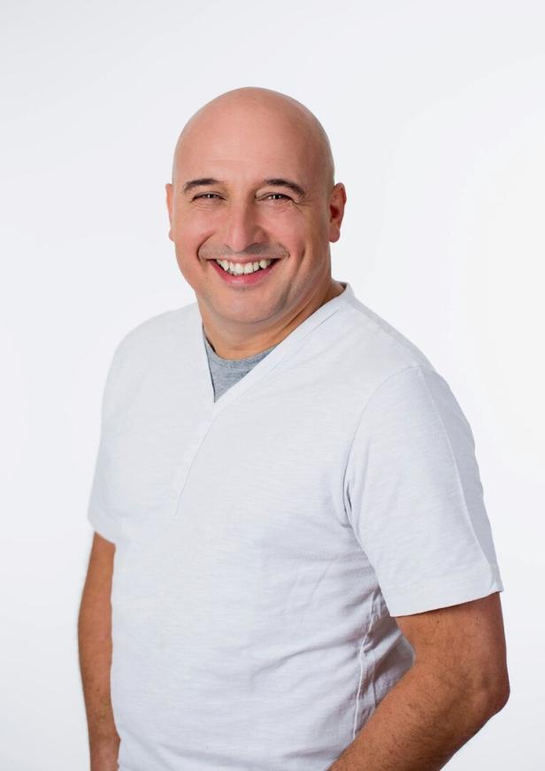 Ein lächelnder, glatzköpfiger Mann in einem weißen T-Shirt vor einem weißen Hintergrund.