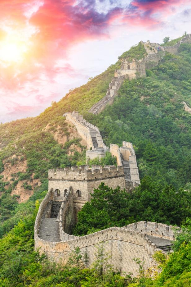 Die Chinesische Mauer schlängelt sich über einen grünen Hügel unter einem farbenprächtigen Himmel.