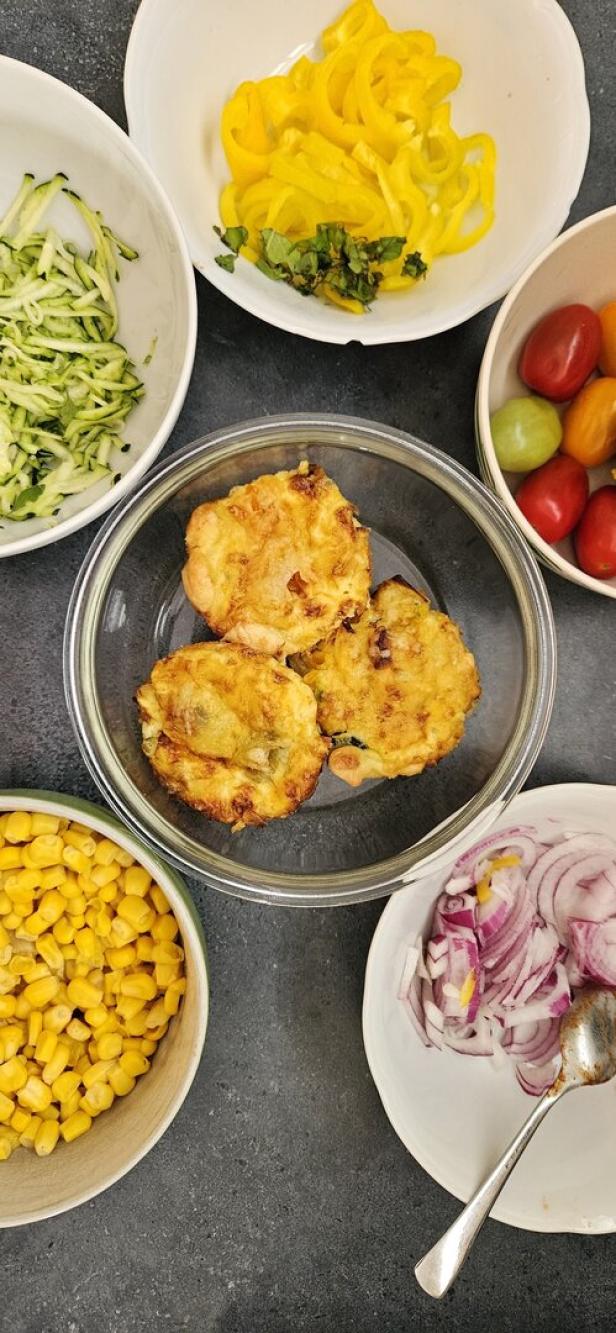 Verschiedene Zutaten wie Mais, Zucchini, Paprika und Zwiebeln umgeben eine Schüssel mit Küchlein.
