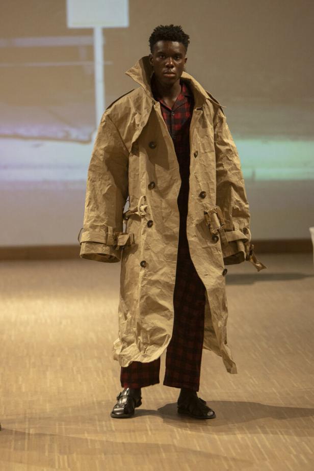 Ein dunkelhäutiger Mann trägt einen beigen Trenchcoat und einen rot-schwarzen Pyjama-Anzug.