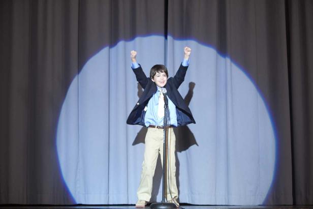 Ein Junge steht auf einer Bühne mit erhobenen Armen im Scheinwerferlicht.