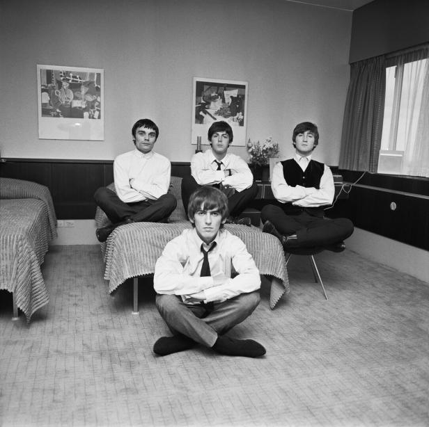 Die Beatles sitzen in einem Hotelzimmer mit gekreuzten Beinen auf Betten und einem Stuhl.