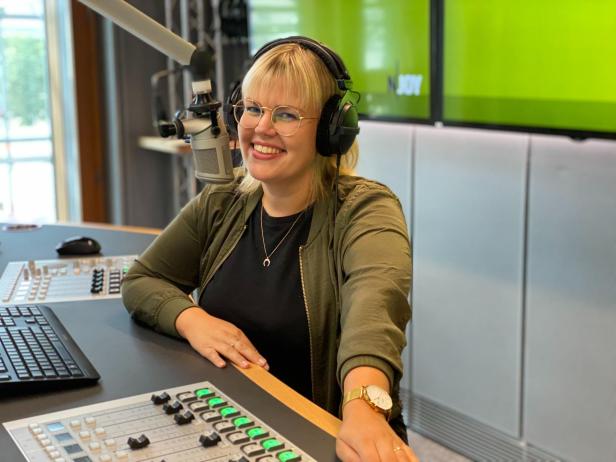Eine lächelnde Frau mit Kopfhörern sitzt vor einem Mischpult in einem Radiostudio.
