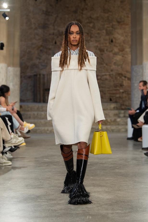 Eine Frau mit Dreadlocks präsentiert ein weißes Kleid, Stiefel mit Fransen und eine gelbe Handtasche auf dem Laufsteg.