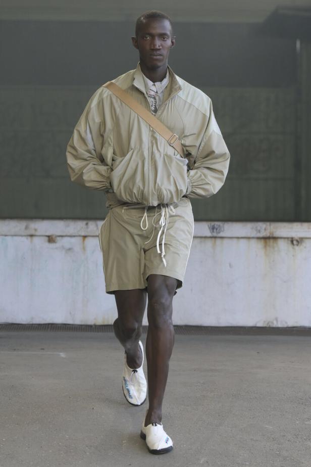 Ein dunkelhäutiger Mann in Shorts und Jacke mit einer Umhängetasche läuft auf einer Betonfläche.