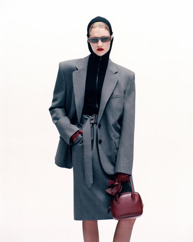 Eine Frau in einem grauen Hosenanzug, Sturmhaube und Sonnenbrille hält eine Handtasche.