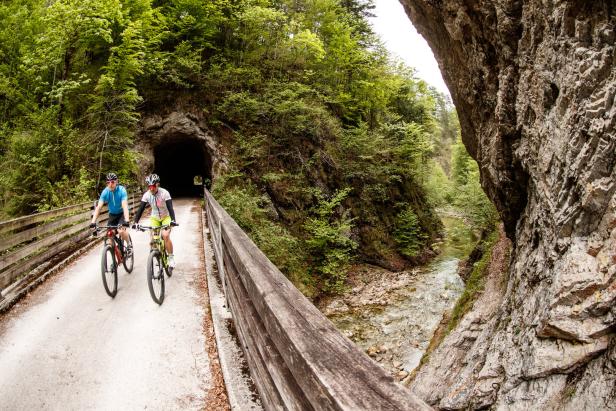 Zwei Radfahrer fahren auf einer Brücke in der Nähe eines Tunnels entlang eines Flusses.