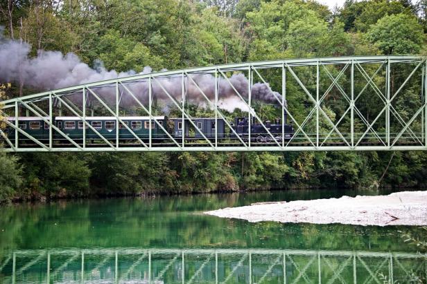 Eine Dampflokomotive fährt über eine grüne Stahlbrücke über einen Fluss.