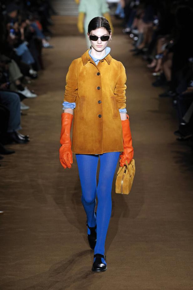 Eine Frau in einem orangefarbenen Wildledermantel, blauen Strumpfhosen und orangefarbenen Handschuhen auf einem Laufsteg.