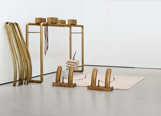 Eine Kunstinstallation mit goldenen Rohren, Holzblöcken und Stoffelementen in einem Ausstellungsraum.