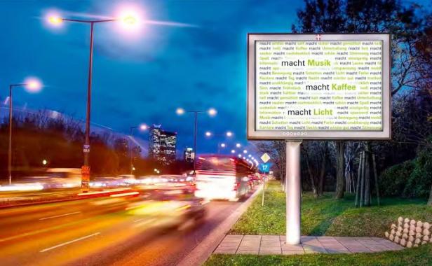 Eine belebte Straße bei Nacht mit einer großen Werbetafel, die mit dem Wort „macht“ und anderen Wörtern gefüllt ist.