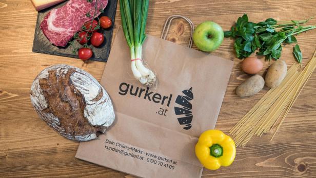 Eine Papiertüte von Gurkerl.at umgeben von verschiedenen Lebensmitteln wie Brot, Fleisch und Gemüse.