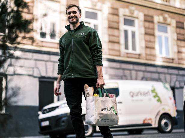 Ein lächelnder Mann mit einer Einkaufstasche von Gurkerl.at vor einem Lieferwagen.