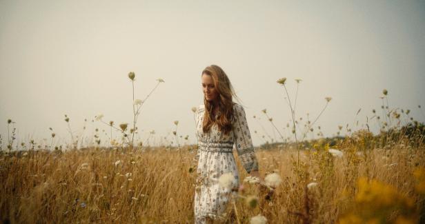 Eine Frau mit langem Haar geht durch ein Feld mit hohem Gras und Wildblumen.