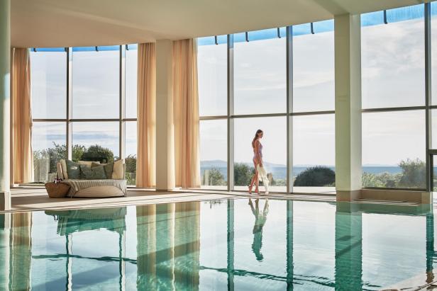 Eine Frau geht in Badebekleidung an einem Innenpool mit Blick auf das Meer entlang.