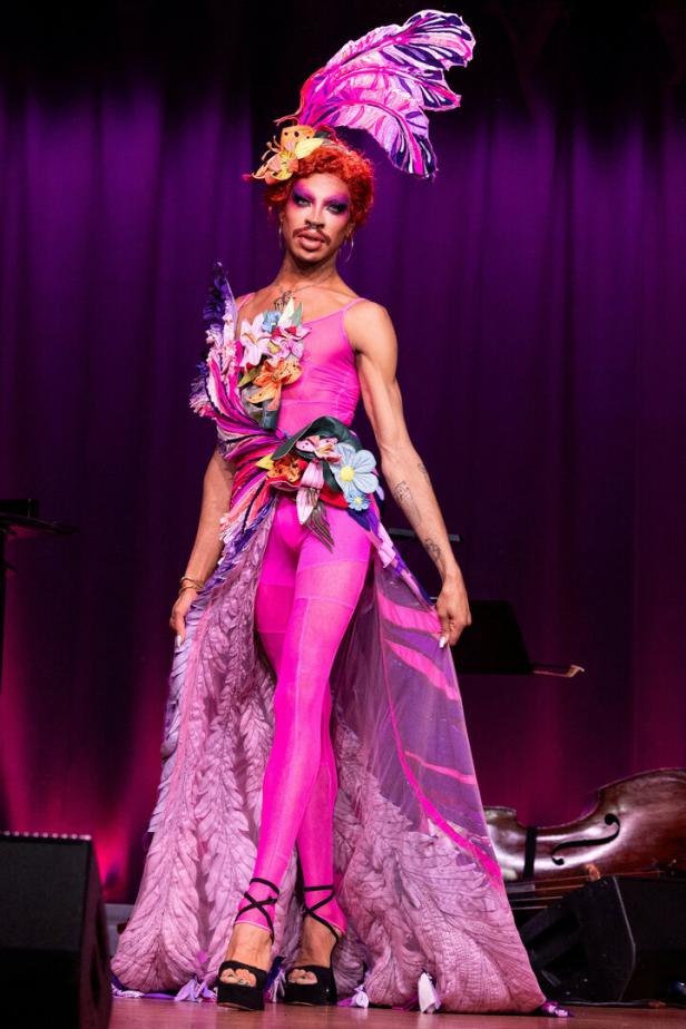 Ein Drag Queen in einem pinkfarbenen Outfit mit Blumen und Federn auf einer Bühne.