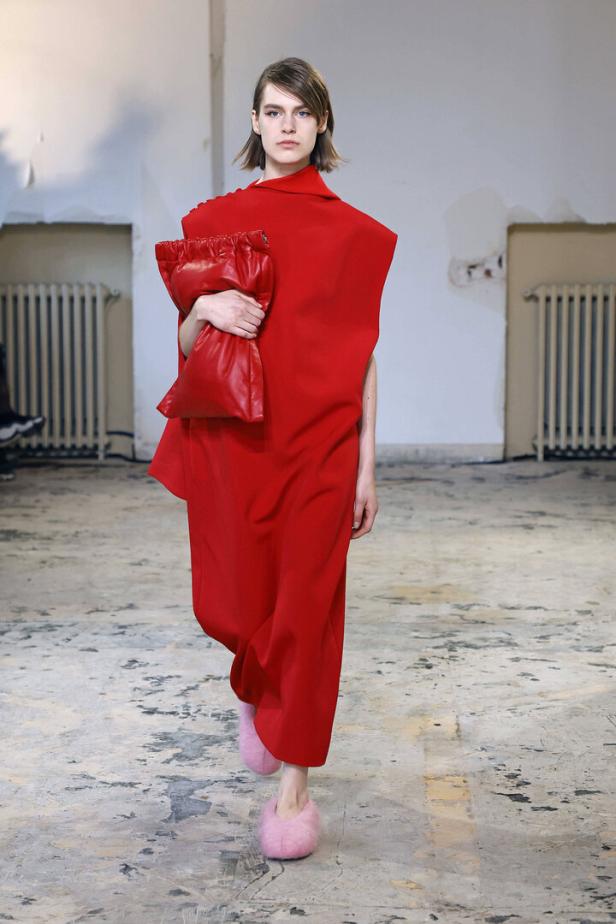 Ein Model in einem roten Kleid und rosa Hausschuhen läuft über einen Laufsteg und hält eine rote Tasche.