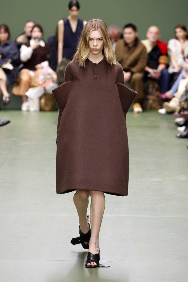 Eine Frau präsentiert ein kastenförmiges, braunes Kleid mit übergroßen Schulterpolstern auf einem Laufsteg.