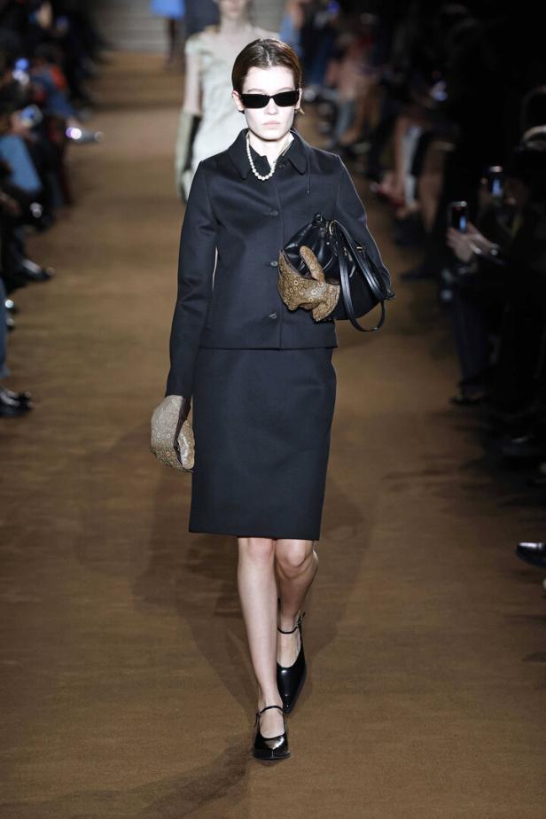 Eine Frau mit Sonnenbrille und Perlenkette trägt ein schwarzes Kostüm und eine Handtasche in Hasenform.