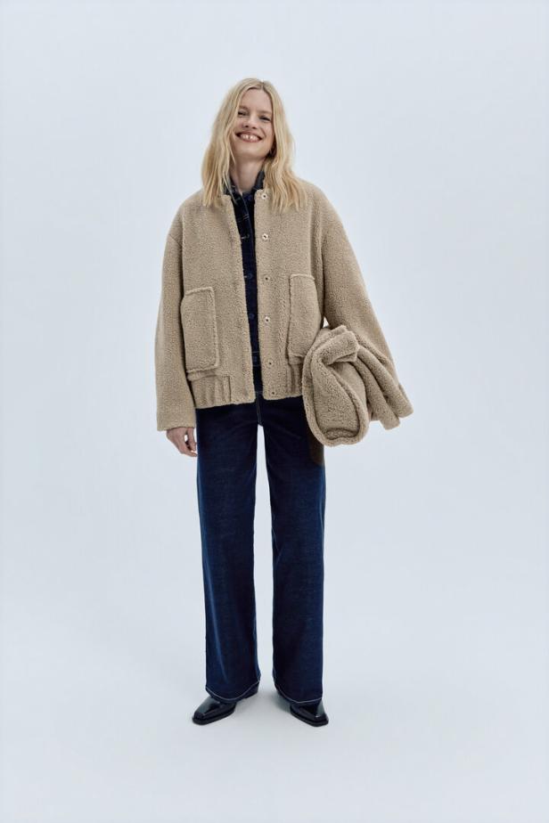 Eine Frau mit blonder Mähne trägt eine Teddyfelljacke und dunkle Jeans.