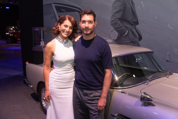 Eine Frau in einem weißen Kleid und ein Mann posieren vor einem Oldtimer.