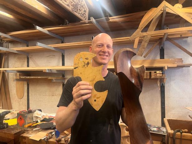 Ein Mann hält eine Schablone und ein bearbeitetes Holzstück in einer Werkstatt hoch.