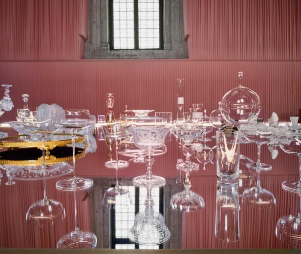 Eine Sammlung von Kristallgläsern steht auf einem spiegelnden Tisch vor einem rosa Vorhang.