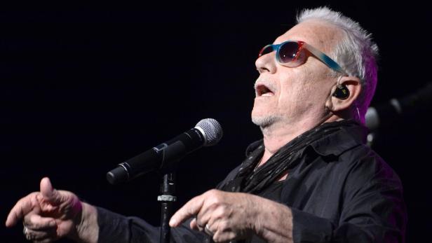 Ein älterer Mann mit Sonnenbrille singt in ein Mikrofon auf einer Bühne.