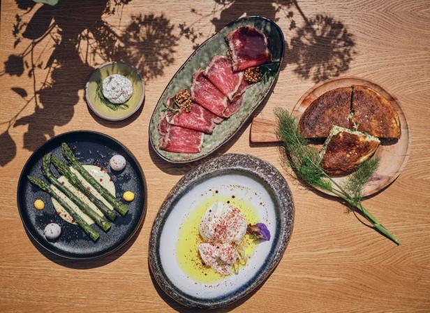 Verschiedene Speisen, darunter Carpaccio, Spargel, Burrata und ein Kräuterbrot, sind auf einem Holztisch angerichtet.