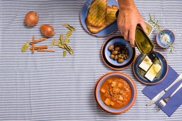 Ein Teller mit Stifado, Oliven, Feta und Brot wird mit Olivenöl beträufelt.