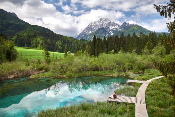Eine Frau sitzt am Ufer eines Bergsees mit Blick auf schneebedeckte Gipfel.