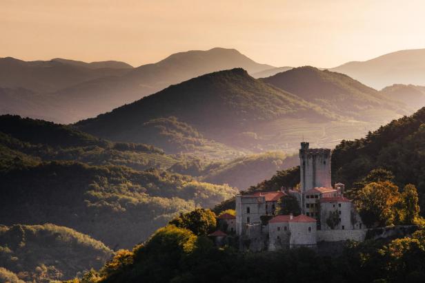 Ein Schloss thront auf einem Hügel, umgeben von bewaldeten Bergen.