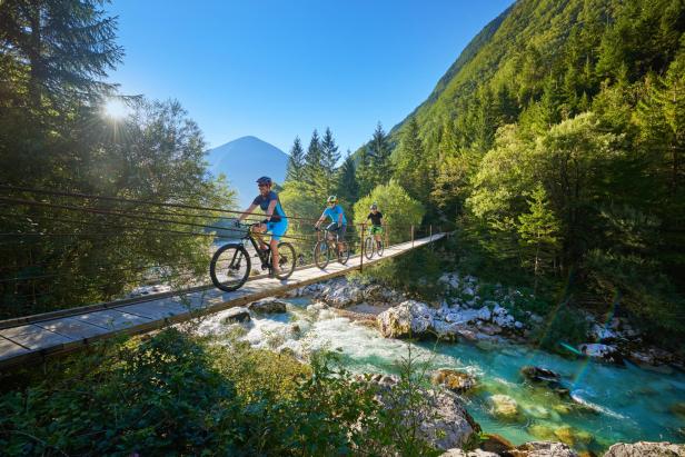 Drei Personen fahren mit Fahrrädern über eine Hängebrücke über einen türkisfarbenen Fluss.