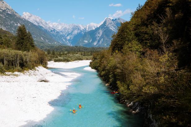 Zwei Kajakfahrer paddeln auf einem türkisfarbenen Fluss vor einer Bergkulisse.