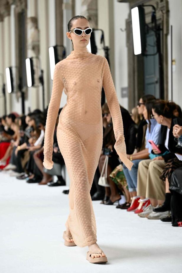 Ein Model mit Sonnenbrille trägt ein transparentes, hellbraunes Kleid auf dem Laufsteg.