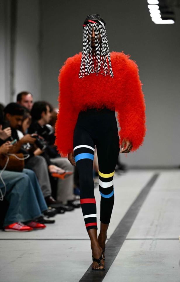 Eine Frau präsentiert ein Outfit mit rotem Felloberteil und Leggings mit bunten Streifen auf dem Laufsteg.