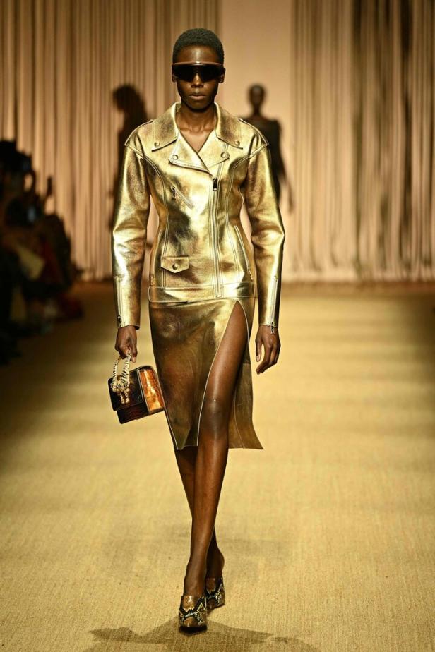 Eine Frau in goldener Jacke und Rock mit Schlitz auf einem Laufsteg.