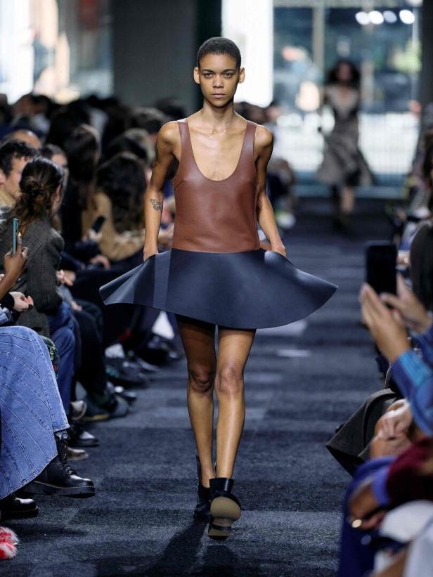Ein Model präsentiert ein braunes Ledertop mit einem ausgestellten, dunklen Rock auf einem Laufsteg.